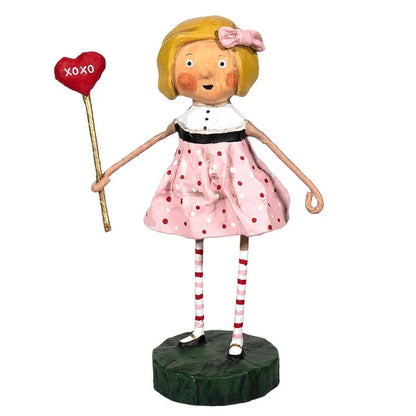 A whimsical figurine of a blonde girl holding a red heart-shaped sign that reads "XOXO." She wears a pink polka-dot dress with a white collar and black waistband, red and white striped socks, and black shoes, standing on a dark green base.