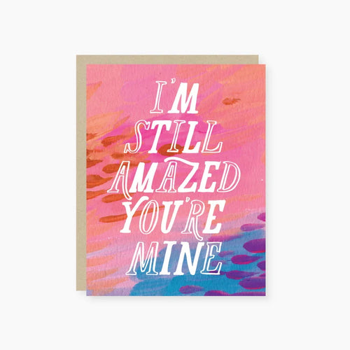 A greeting card with a colorful background and the text 'I'm Still Amazed You're Mine' in bold letters. The card also has a matching envelope.